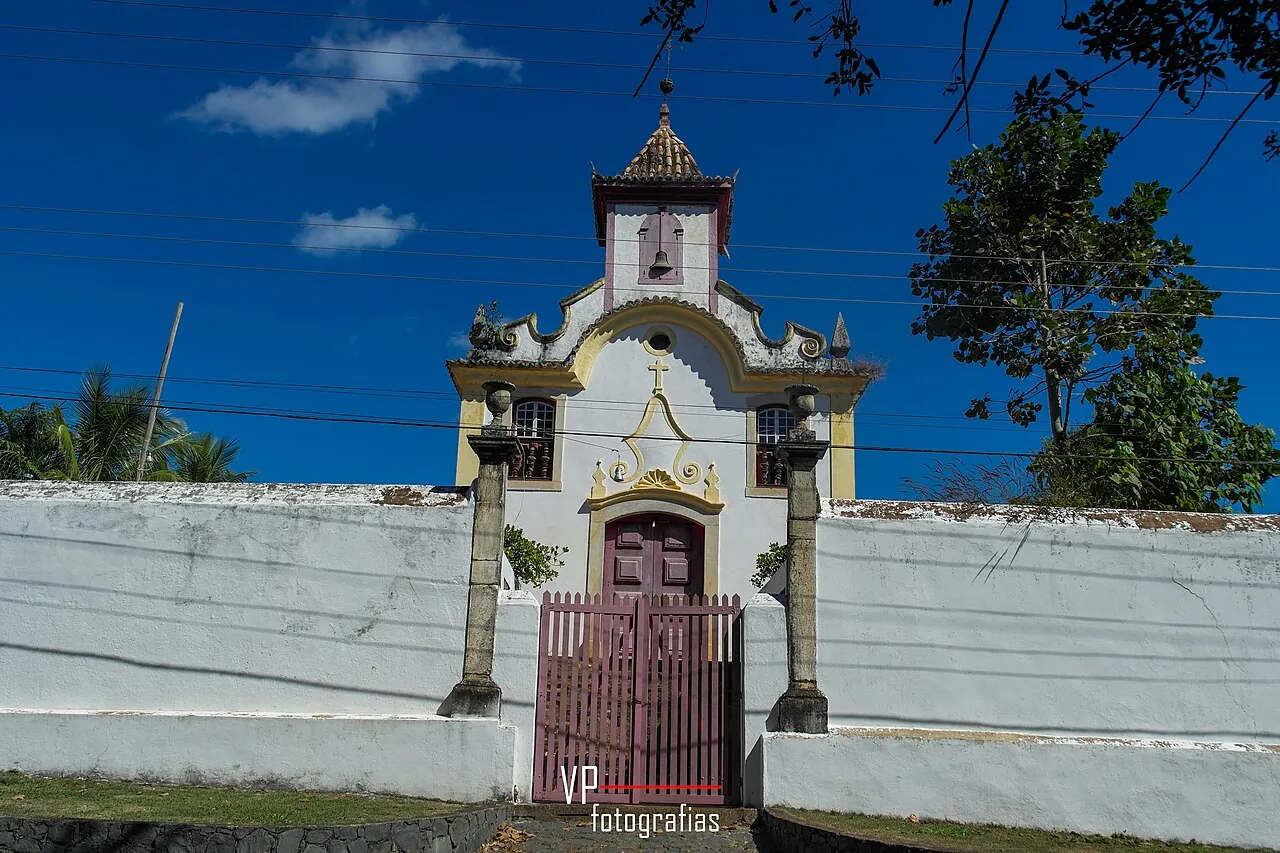 Igreja histórica de Cocais com fachada branca, frontão em volutas amarelas, torre sineira central e portão vinho no muro caiado, sob céu azul, em Barão de Cocais (MG)